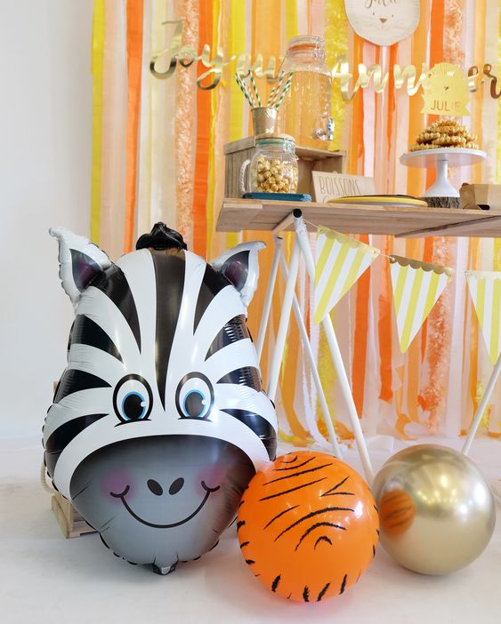 A party decor set up including a foldable table, several snack items on the table, and three balloons positioned under the table including a zebra bust mylar, a tiger fur patterned latex, and a chrome gold latex.