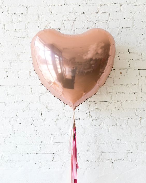 Single, jumbo sized rose gold balloon with a pink tassel, set against a white brick wall